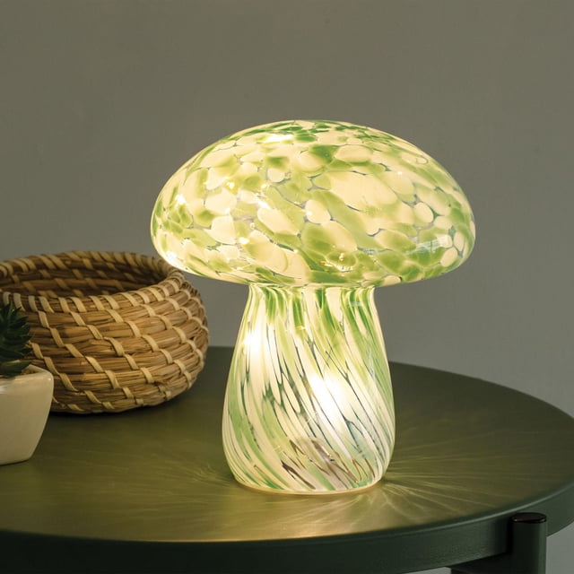Merkury Innovations 7.9 inch Height Green Confetti Glass Mushroom Lamp with a mushroom-shaped glass shade featuring green and cream confetti patterns and a swirled glass base, casting a soft warm glow on a green table beside a woven basket and small plant, suitable for ambient decorative lighting in home interiors.