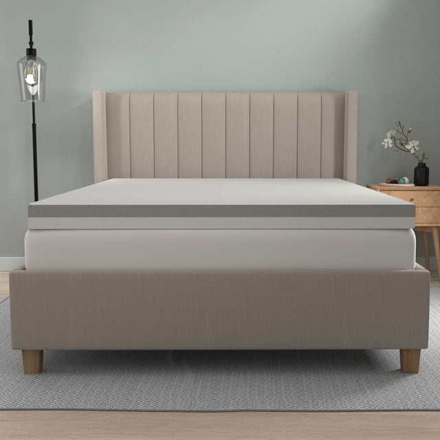 Dream Serenity® 4-inch Slab Mattress Topper - Q shown on a queen bed with a beige upholstered headboard featuring vertical channels. The topper has a light grey top and darker grey side panel, resting on a white mattress within a beige fabric bed frame with wooden legs. The bed is on a grey patterned rug, flanked by a black floor lamp and a wooden nightstand with a black vase and white flowers against a pale blue wall. The topper is also suitable for adding extra cushioning to guest beds or daybeds.