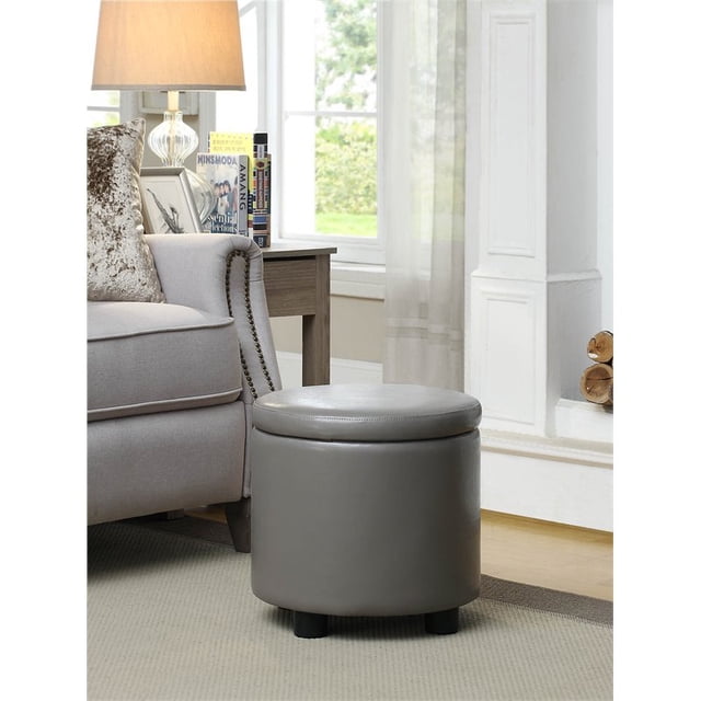 Designs4Comfort Round Accent Storage Ottoman in gray faux leather fabric, featuring a compact cylindrical design with a removable lid that doubles as a multifunctional tray. The ottoman is placed in a cozy living room setting next to a beige armchair and a wooden side table. It offers concealed storage and measures 16 inches in height and 15.75 inches in diameter.