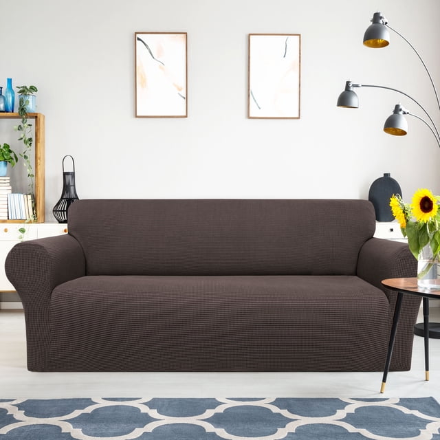 PiccoCasa Housse de Sofa Extensible en Brun Chocolat, Conçue pour les sofas de Taille Moyenne. Fabriquée à Partir de 95 % de polyester et 5 % d'Élasthanne, Cette Housse de Meuble Lavable Est Douce, Respirante et Très Extensible pour un Ajustement Serré. la Housse de Sofa Présente des Bandes Élastiques et des Bandes de Mousse pour la Maintenir Bien en place, Rajeunissant l'Apparence de Votre Sofa Tout en le Protégeant de l'Usure. Exposée dans un Décor de Salon Moderne avec une Déco Neutre et un Tapis à Motifs.