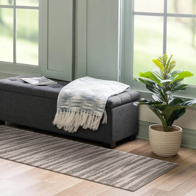 Tapis Coureur AVB Vague Verte 2x7 avec Rayures Texturées Ton Terre sur Plancher de Bois Clair, Posé à Côté d'Un Banc Capitonné Gris Foncé avec un Jeté à Franges et une Plante Verte en Pot Près de Grandes Fenêtres, Idéal pour les Couloirs Ou les Entrées.
