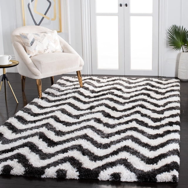 Safavieh Barcelona Carmella Zigzag Stripes Shag Area Rug with plush black and white zigzag stripes on dark hardwood floors. Displayed in a bright room with a beige accent chair, patterned pillow, small side table with a white cup, and a white vase with green palm leaves near French doors. Ideal for adding texture and style to accent rug spaces.