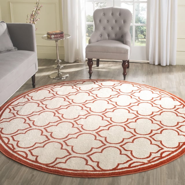 Safavieh Amherst Wilson Tapis Géométrique pour Aire, Rond avec une base Blanche et Motif Trèfle Rouille, Polypropylène Tissé Mécaniquement pour une Utilisation durable Intérieur-Extérieur, Montré sur un Plancher de Bois Clair dans une Pièce Lumineuse avec un Fauteuil Capitonné Gris, un sofa Gris et une Petite table d'Appoint Ronde en Métal avec des Livres et des Fleurs.