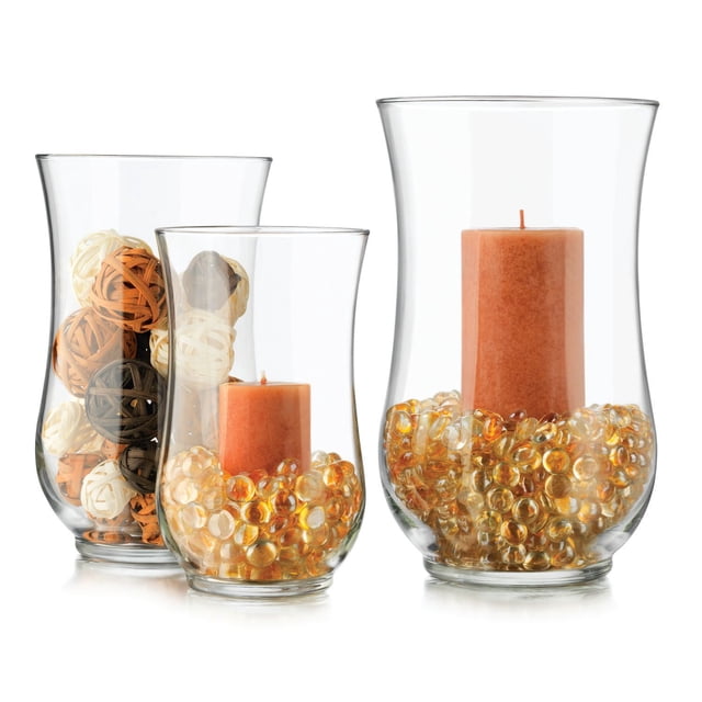 Angela Ensemble Ouragan de Trois vases Ouragan en Verre Libbey Transparents avec Hauts Évasés; Deux Contiennent des Bougies Piliers Ambrées Entourées de Perles Décoratives Ambrées Translucides, et l'Un Contient des Boules en Rotin Tressé Ton Naturel, Convient pour Rehausser la Déco de Table Ou des Événements Spéciaux.