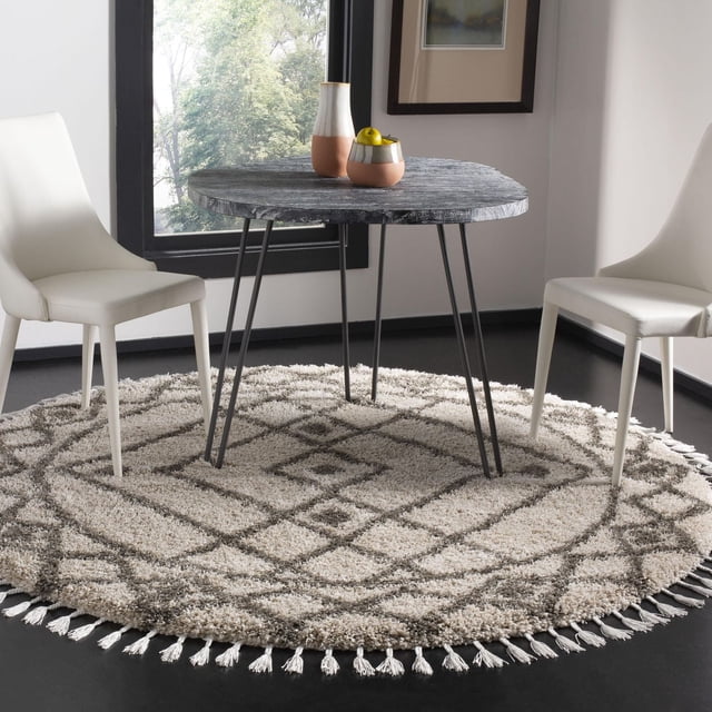 Safavieh Moroccan Fringe Erksine Geometric Shag Area Rug in round shape with beige and gray tribal geometric pattern and fringed edges, placed under a dark wood round table with black metal hairpin legs and two white modern dining chairs, suitable for adding plush texture to dining areas.