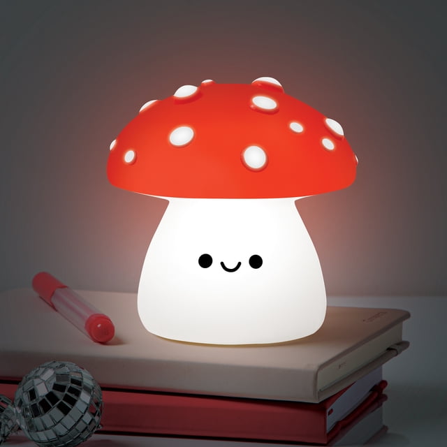Merkury Innovations Veilleuse LED Champignon Magique en Blanc Froid, en Forme de Champignon Souriant avec une Calotte Rouge et des Taches Blanches, Projetant une Lueur Douce sur des Livres Empilés avec un Stylo et une Boule disco à Proximité, Idéale pour des Accessoires d'Éclairage Intérieur et Créant une Atmosphère Apaisante au Coucher.