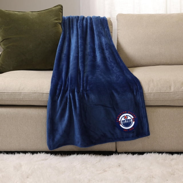 NHL Blue Sports Throw Blanket, 50" x 60", plush deep blue polyester blanket draped over a beige sofa with a green cushion, featuring an embroidered Edmonton Oilers logo patch, suitable for cozy indoor or outdoor use.