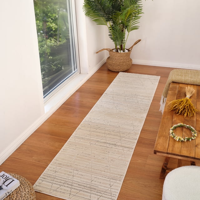 Rug Branch Astra Modern Indoor Area Rug, Beige Cream, Abstract, displayed as a runner on a hardwood floor in a bright room with natural light. The polyester rug with jute backing features a geometric pattern and 0.4-inch pile height, ideal for hallways and entryways. Positioned near a large window beside a woven basket with a green plant and a wooden bench with dried flowers and a floral wreath, it offers durable floor protection and a fresh look.