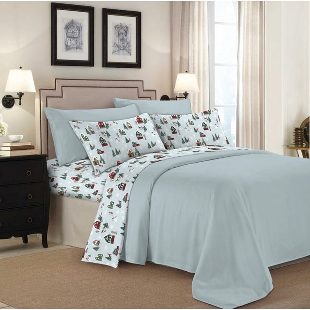 Johnson Home 144 Thread Count Cotton Printed Sheet Sets on a bed with beige upholstered headboard, featuring festive holiday-themed percale cotton sheets with small houses and trees in red, green, and brown on white. Includes deep 15-inch pocket fitted sheet and solid light blue pillowcases and top sheet. The bedroom has black nightstands with lamps and framed architectural prints.
