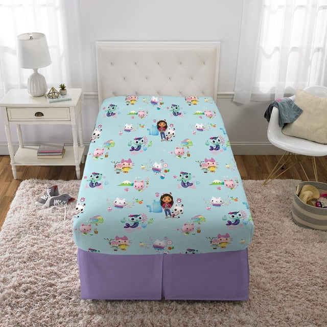 Gabby's Dollhouse "Paw Tastic" twin sheet set featuring a light blue fitted sheet with colorful cat-themed characters from the DreamWorks series, on a twin bed with a white tufted headboard in a children's bedroom with white walls, sheer curtains, a white nightstand with a lamp, a white chair with a cushion, and a beige rug on hardwood flooring.