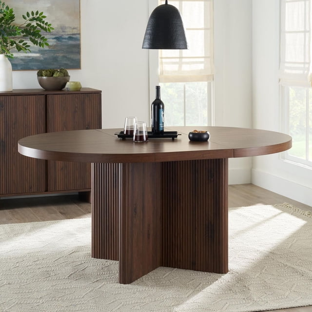 Better Homes & Gardens Lillian Fluted Round Expandable Dining Table in walnut finish with circular tabletop and fluted crossed base, set on a beige textured rug in a bright dining room with a black pendant light and matching walnut sideboard, ideal for various room sizes.