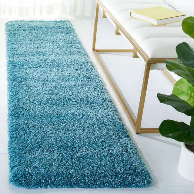 Safavieh Milan Harlow Solid Shag Area Rug in aqua blue, a rectangular synthetic shag runner on a white floor beside a white cushioned bench with gold metal legs and a yellow book, near a green potted plant, suitable for adding texture and style to modern home interiors.