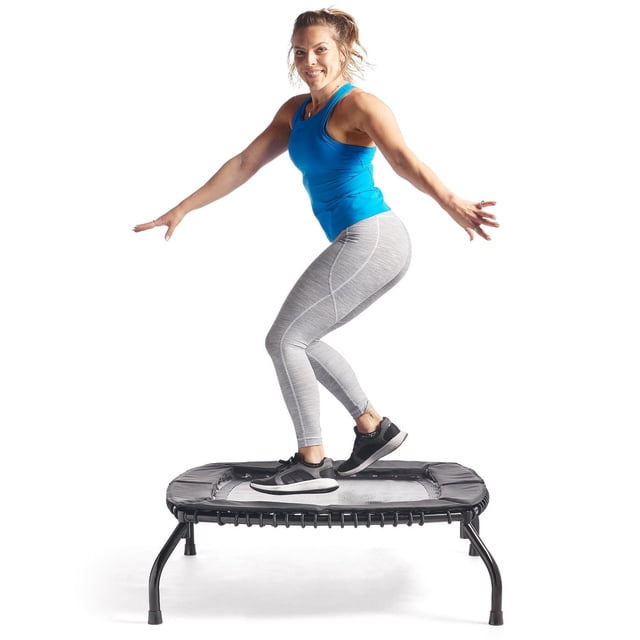 Sunny Health & Fitness Springless Mini Fitness Trampoline with a woman balancing on its square frame featuring adjustable black bungee cords and sturdy legs, wearing a blue tank top, grey leggings, and black sneakers on a white background.