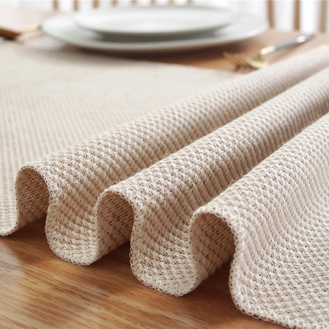 Chemin de Table Style Ferme, Chemin de Table Rustique Vintage, Chemin de Table Basse Tressé en Coton beige, Montré en Gros Plan Mettant en Valeur Son Épaisse texture en Coton Tissé avec de Délicats Glands aux Deux Extrémités. le Chemin de Table Mesure 33x180 cm (72 Pouces de long) et Présente un design Naturel et Rustique Convenant à une Déco de Maison de Style Campagnard. Il Est Exposé sur une Table à Manger en Bois avec des Assiettes et des Couverts en Arrière-Plan.
