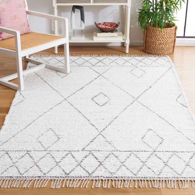 SAFAVIEH Casablanca Abby Bohemian Area Rug in white with a hand-tufted New Zealand wool shag texture, geometric diamond pattern, and fringed edges on a light hardwood floor beside a white wooden chair with woven seat and pink cushion, near a white console table and woven basket plant holder.