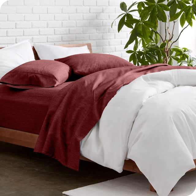 Bare Home Premium 1800 Collection 4-Piece Bed Sheet Set for Queen beds in Heathered Rose color, shown on a wooden bed frame with white bedding accents. This luxury bedding set includes a fitted sheet, flat sheet, and two pillowcases crafted from ultra-soft, hypoallergenic microfiber with a 1800 thread count. The fitted sheet features deep pockets to fit mattresses up to 15 inches deep. The minimalist bedroom setting includes a white brick wall and a green potted plant for added decor.