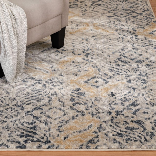 Pescara Blue and Grey Woven Area Rug with a faded vintage ornate pattern in charcoal, dusty grey, and gold tones, shown under a beige sofa leg with a knitted throw, made of stain-resistant polypropylene with a low profile pile suitable for living rooms and bedrooms.