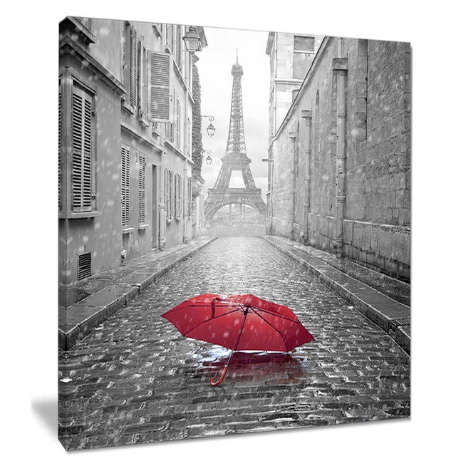 Design Art Eiffel View from Paris Street Cityscape Photo Canvas Art Print featuring a gallery wrapped canvas wall art of a rainy Paris cobblestone street with a vivid red umbrella lying open on the wet pavement, the Eiffel Tower framed by classic Parisian buildings in the misty distance, ready to hang with included kits and suitable for enhancing home or office décor.