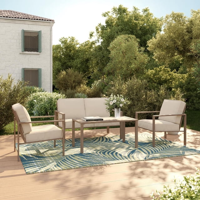 Mainstays Cobble Hill 4-Piece Steel Outdoor Conversation Set with Cushions, Cream, featuring a loveseat and two armchairs with UV- and fade-resistant cream cushions on rust-resistant, powder-coated steel frames with hand-painted woodgrain finish, arranged around a rectangular steel slat tabletop on a wooden patio deck with a blue and green leaf-patterned outdoor rug, set against green shrubbery and a stone house with green shutters, ideal for outdoor dining and relaxing.