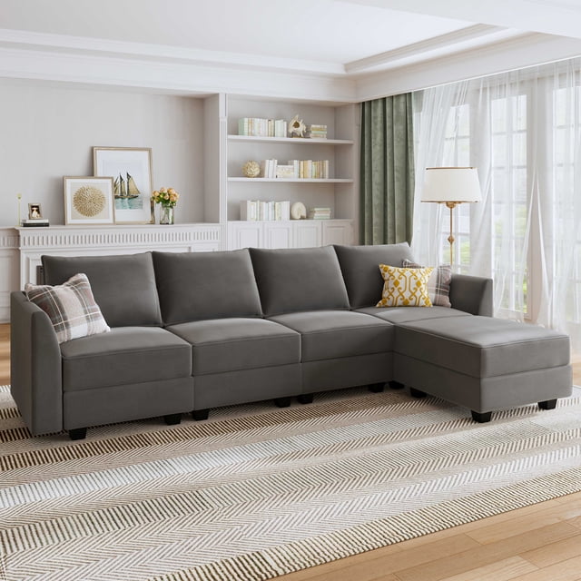 HONBAY Velvet Couch with Reversible Chaise and Storage Ottoman in dull grey, displayed in a bright living room setting. The sectional sofa features a modular design with soft velvet upholstery, offering a luxurious and comfortable seating experience. The reversible chaise allows for flexible arrangement, while the storage ottoman provides a practical compartment for magazines, books, or electronics. The dull grey color complements the neutral decor of the room, which includes a patterned rug, a floor lamp, and built-in shelves.