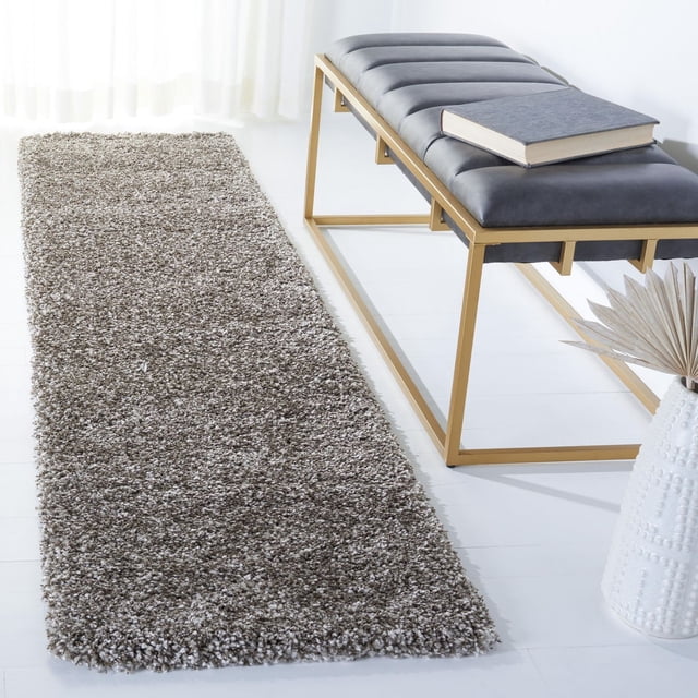 Safavieh Milan Harlow Tapis Shag Uni Gris, un Coureur en Fil Synthétique Pelucheux à texture Shag dense, Posé sur un Plancher Blanc à Côté d'Un Banc Moderne à Cadre Métallique Doré avec Siège Rembourré Gris Foncé et un Livre sur le Dessus, Accompagné d'Un vase Texturé Blanc avec des Feuilles de Palmier Séchées, Convient pour Ajouter Chaleur et style à la Déco Contemporaine de la Maison.