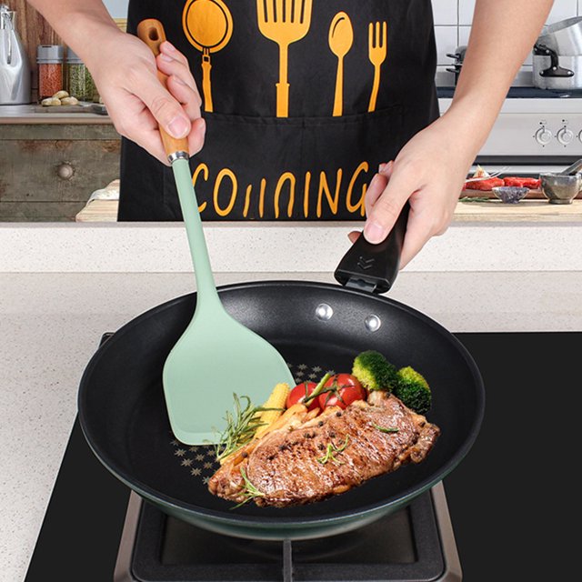 TIMIFIS Steel Spatula with an exclusive wooden handle and a green non-stick silicone head, shown in use flipping a steak in a frying pan alongside vegetables. The spatula is lightweight, food-grade, and designed for versatile kitchen use, including frying, baking, and outdoor cooking. The wooden handle provides a comfortable grip, and the silicone head is flexible for easy scraping and cleaning.