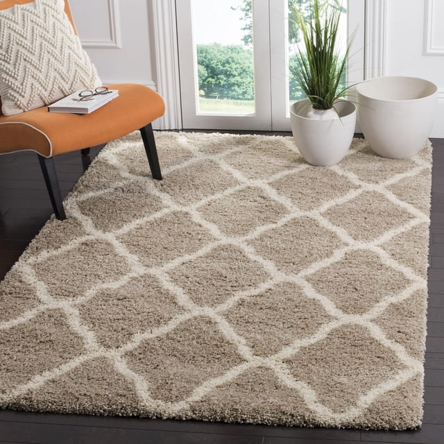 Safavieh Hudson Aline Geometric Shag Area Rug with beige and white Moroccan tile pattern on dark hardwood floor, shown in a bright room with an orange chair featuring a cream textured pillow and two white ceramic planters, one holding a tall green plant, suitable for enhancing comfort and décor.