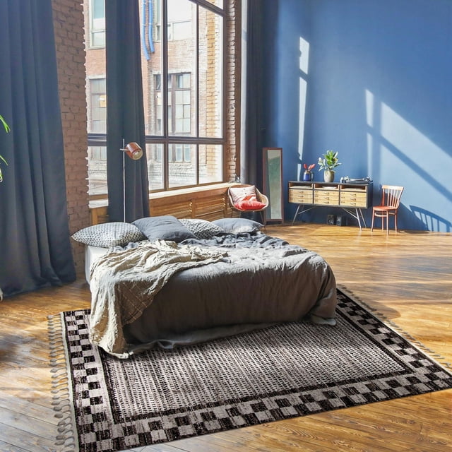 ECARPET Southwestern Rug for Hallway, Kitchen Quattro Collection in grey with a checkered border and woven texture, placed under a low bed with grey bedding in a sunlit room featuring wooden floors, exposed brick walls, and large industrial windows, ideal for high-traffic indoor spaces.