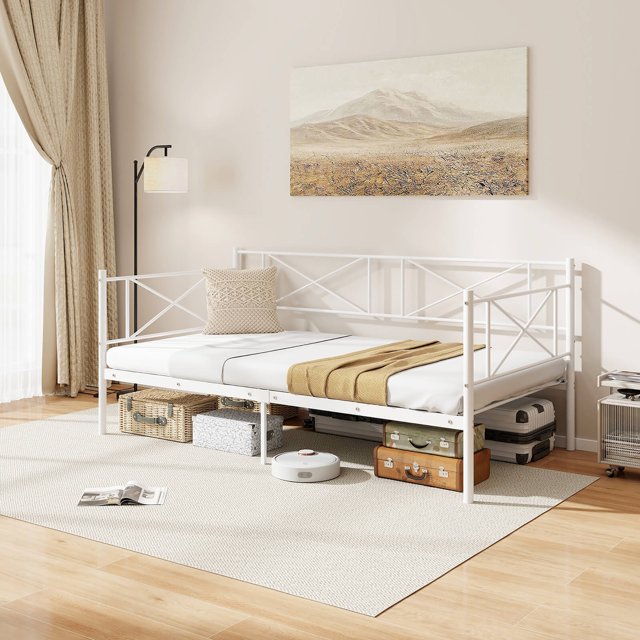 Gymax Twin Size Metal Daybed in white with a modern platform design featuring heavy-duty metal slat support and a cross-shaped decorative backrest and armrests. The daybed is shown in a bright, minimalist room with wooden flooring and a beige rug. It includes ample under-bed storage with visible suitcases and boxes. The bed frame supports up to 485 lbs and does not require a box spring, making it ideal for small spaces or guest rooms.