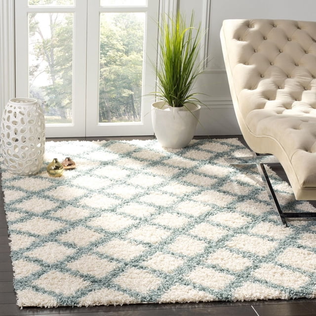 Safavieh Dallas Jerrie Geometric Shag Area Rug featuring a plush diamond pattern in soft white and light blue synthetic fibers, placed on dark hardwood flooring beside a beige tufted sofa and a white ceramic planter with green plant, suitable for high-traffic home areas.