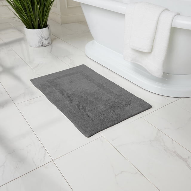 Fabstyles Basix Tapis de Bain en Coton Réversible Doux et Absorbant Gris, Placé sur un Plancher de Salle de Bain à Carreaux de Marbre Blanc, à Côté d'Une Baignoire Autoportante Blanche avec une Serviette Blanche Pliée au-Dessus du Bord et une Plante Verte dans un pot en Marbre, Convient pour Améliorer le Confort et l'Absorption de la Salle de Bain.