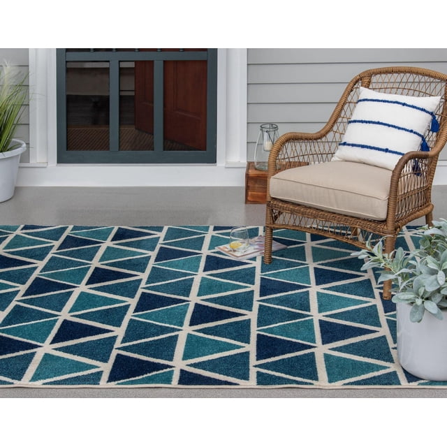 Tropiana Cream and Turquoise Blue Woven Area Rug with geometric triangle pattern in sapphire, turquoise, and ivory on a patio floor next to a wicker chair with beige cushion and white pillow with blue stripes, accompanied by potted plants and a glass of lemon water on a magazine, ideal for indoor or outdoor spaces.