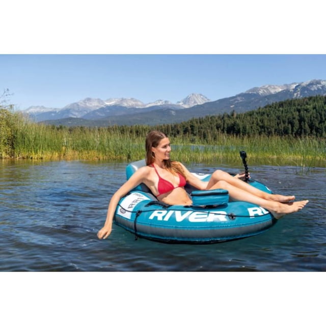 Intex River Run 1 Pro single rider inflatable water raft in blue with white 'RIVER RUN' text, featuring a backrest, cup holder, and grab rope, floating on a calm lake surrounded by green reeds and forested hills with snow-capped mountains in the distance under a clear blue sky, with a person relaxing in a red bikini and an action camera mounted on the raft, ideal for leisurely floating on rivers or lakes.