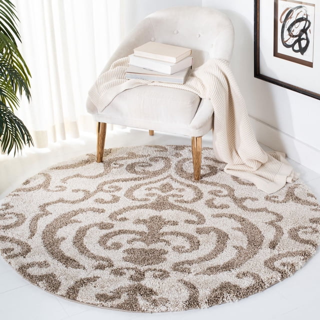 Safavieh Florida Deangelo Damask Plush Shag Area Rug, round with a plush high-low pile damask pattern in crème tones, placed under a cream upholstered chair with wooden legs, featuring a beige knit throw and stacked books in a bright room with white walls and framed abstract art, suitable for adding soft texture to living spaces.