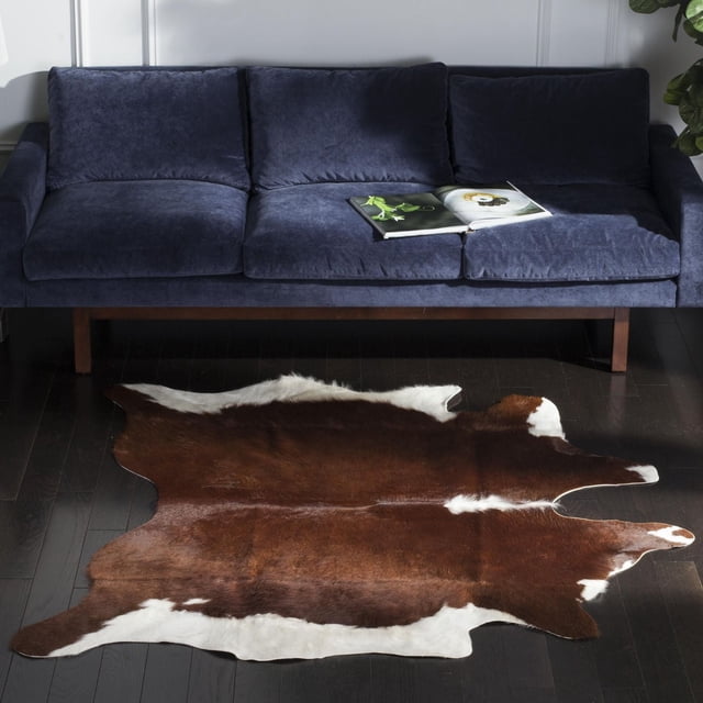 Safavieh Cow Hide Donovan Tapis Animal sur Plancher de Bois Franc Foncé devant un sofa Trois Places Bleu Marine avec Pattes en Bois. le Tapis Présente une Peau de Vache Brun Naturel et Blanc avec des Bords Irréguliers et une texture visible, Idéal pour Ajouter un style Rustique et une Durabilité aux Salons.