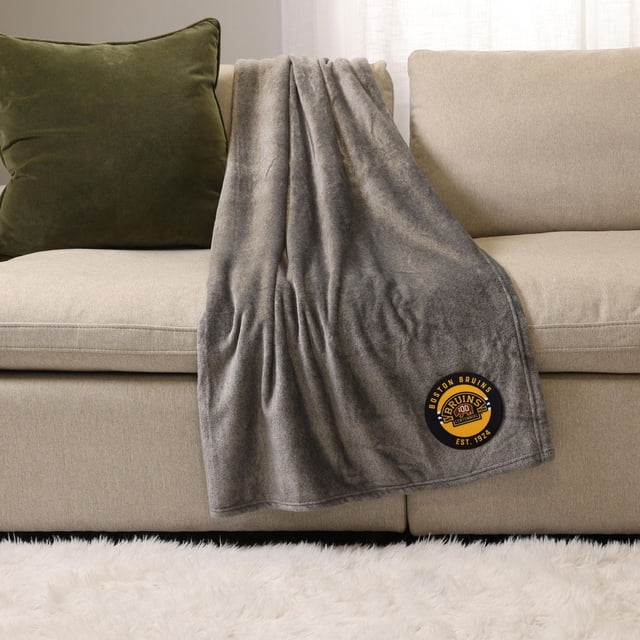 NHL, NBA, MLB Grey Sports Throw Blanket, 50" x 60", draped over a beige sofa with a green cushion, featuring a Boston Bruins logo patch in black and gold near the bottom corner. This plush 100% polyester throw blanket provides cozy warmth for indoor or outdoor use.