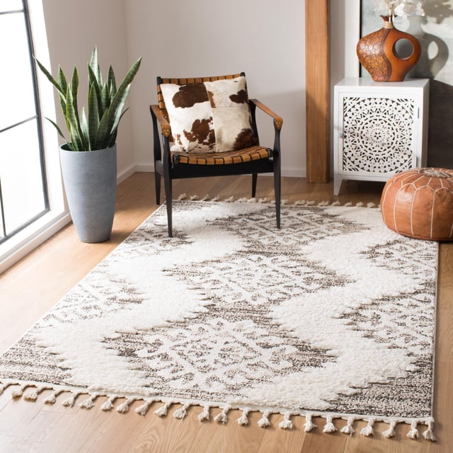 Safavieh Moroccan Tassel Shag Desmond Southwestern area rug in cream and brown geometric pattern on wooden floor next to black chair with cowhide cushion, tall green plant in gray pot, white carved cabinet topped with terracotta vase and leather pouf.