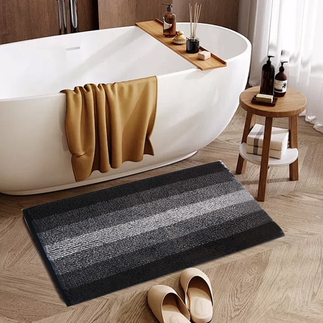 TIMIFIS Bathroom Rugs Absorbent Entrance Mat, a modern striped bathroom rug in black and grey tones, designed for water absorption and non-slip functionality. Positioned near a freestanding bathtub in a contemporary bathroom with wooden flooring and decor accents like a stool and bath accessories. Made from 95% polyester and 5% spandex, this bath mat measures 40x60cm and is ideal for bathrooms, shower rooms, and toilets.