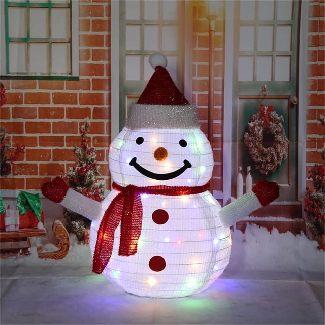 Snowman Outdoor Christmas Decorations Light Christmas Decoration Outdoor Christmas Lighting Outdoor Figures, Santa Claus Snowman LED with 40 LEDs Foldable. The image features a smiling snowman with a red scarf, red buttons, and a red Santa hat, illuminated by 40 LED lights. The snowman is displayed outdoors against a festive backdrop with a brick wall, wreath-adorned doors, and snow-covered ground. This foldable decoration is easy to install and store, perfect for creating a festive atmosphere in gardens, driveways, or front yards.