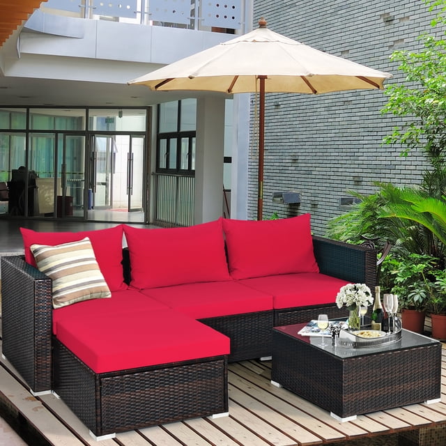Gymax Ensemble de Conversation de Patio en Rotin Rembourré 5 Pcs avec Ottoman Comprenant des Coussins Rouges, Incluant 2 Chaises d'Angle, 1 Chaise Sans Accoudoirs, 1 Ottoman et 1 table Basse avec Dessus en Verre Trempé. Idéal pour une Utilisation Extérieure, Tels Que les patios, les Jardins Ou les Zones au Bord de la Piscine, l'Ensemble Est Exposé sur une Terrasse en Bois Entourée de Verdure et un Parasol de Patio beige.