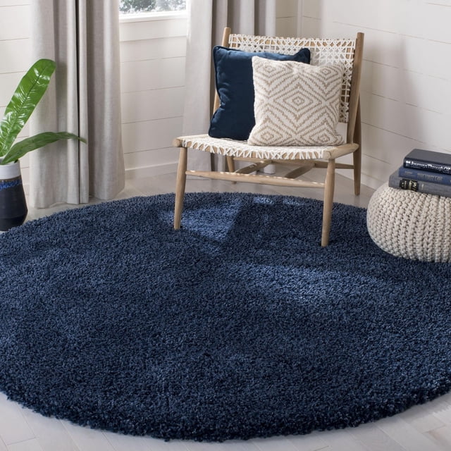 Safavieh California Tapis Shag en Peluche Uni, Rond, Bleu Marine Foncé avec un Empilage de Polypropylène dense de 4,5 Kg/m2, Posé sur un Plancher de Bois Pâle, à Côté d'Une Chaise en Bois Tressée avec des Coussins Bleu Marine et beige, un pouf Tricoté beige et une Plante Verte Près de Rideaux beige et d'Une Fenêtre. Ce Tapis Pelucheux Est Souvent Utilisé pour Améliorer des Espaces de Détente Ou de Lecture Douillets.