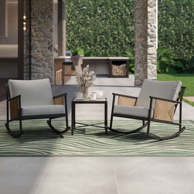 HOMETRENDS 3-Piece Steel Outdoor Rocking Chair Chat Set with Table & Cushions, Tan, featuring two steel-framed rocking chairs with tan all-weather resin wicker sides and gray UV- and fade-resistant cushions, paired with a durable steel table with a stone-like top, set on a green patterned outdoor rug on a patio with stone pillars and greenery, ideal for outdoor conversation and relaxation.