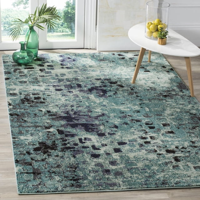 Safavieh Monaco Driskoll Abstract Area Rug with power-loomed polypropylene texture in teal, navy, and black abstract patterns, placed on light wood flooring under a white triangular coffee table with wooden legs holding a glass vase of lemons and a small succulent, near green glass vases and palm leaves by a window. Suitable for adding durable, Bohemian-chic accent to home interiors.