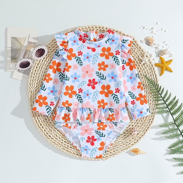 Toddler Girl Rash Guard Swimming Bathing Suit with long sleeves and a vibrant floral pattern in orange, pink, and blue tones. Designed for sun protection, this bathing suit features ruffle detailing at the waist and is suitable for ages 2-3 years. Displayed on a woven mat with accessories like sunglasses, seashells, and a starfish, evoking a summer beach vibe.