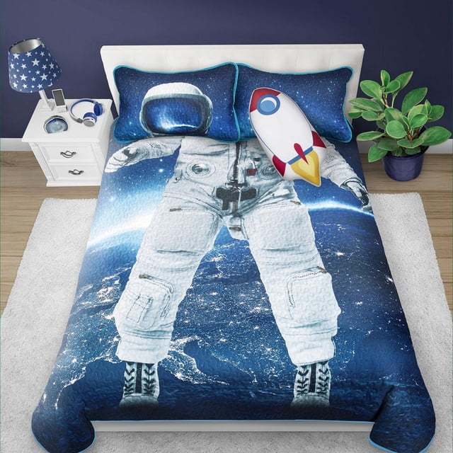 Safdie & Co. Quilt 3PC Set DQ Astronaut with a quilt showing an astronaut in a white suit standing over a nighttime Earth view, two blue starry pillow shams, and a rocket-shaped pillow on a white bed frame. Made of soft, breathable polyester microfiber, it adds comfort and space-themed style to any bedroom.