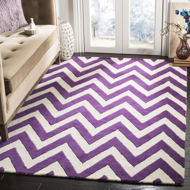 Safavieh Cambridge Kaitlyn Zig Zag Stripes Area Rug in purple and white hand-tufted wool with a zigzag pattern, placed on dark hardwood flooring beside a beige tufted sofa and decorative accents, suitable for enhancing modern home or office decor.