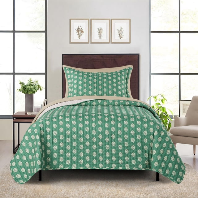 Mainstays Quilt-in-a-bag bedding set featuring a green quilt and matching pillow sham with white floral patterns and beige trim on a bed with a dark wood headboard. The set includes a lightweight quilt and pillow sham made from recycled polyester. The bedroom has large windows, a side table with a plant and books, three framed botanical prints above the bed, and a beige armchair. This bedding set is ideal for adding comfort and style to any bedroom.