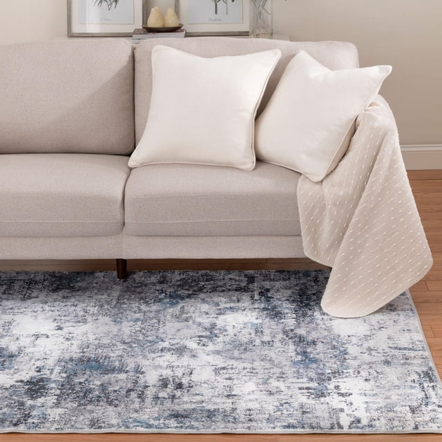 Banburry Ocean Blue and Grey Woven Area Rug with an abstract distressed pattern in charcoal, slate blue, cobalt, and off white, placed on a hardwood floor under a beige sofa with two off-white pillows and a beige throw. Suitable for adding modern rustic style and softness to living rooms or cozy spaces.