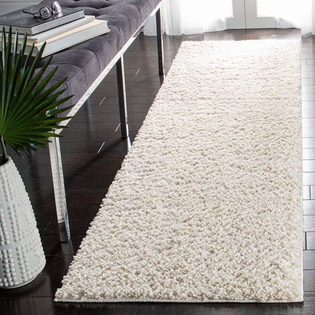 SAFAVIEH August Carlene Solid Plush Shag Area Rug in off-white, displayed on dark hardwood flooring next to a gray tufted bench with chrome legs, featuring a thick plush polypropylene yarn texture suitable as an accent rug in modern home interiors.