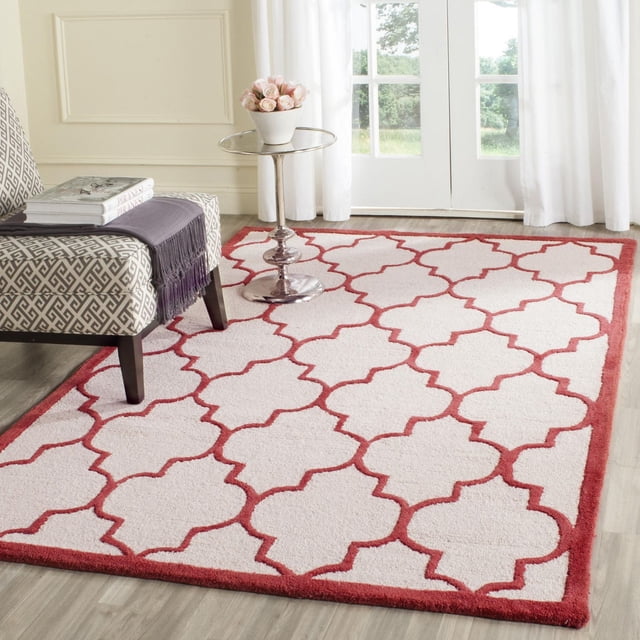 Safavieh Cambridge Kevin Geometric Area Rug with hand-tufted wool construction, white base, and red geometric trellis pattern, placed on light hardwood floor beside a grey patterned armchair and a silver round side table with a white vase of pink roses, near white curtains and glass doors providing natural light.